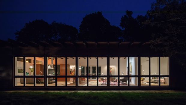 Night-time exterior view of a low, elongated modern residence with extensive floor-to-ceiling glazing and dark window frames. Warm interior lighting reveals an open-plan living area, kitchen, dining space and furnishings behind the glass. A dark horizontal roof/pergola element and surrounding mature trees frame the facade.