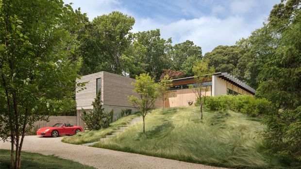 A contemporary low-profile house set into a lush, heavily wooded landscape. The exterior combines horizontal timber cladding, pale rendered or concrete walls, warm timber panels, and a dark roof edge. A stepped path crosses a gently sloping lawn planted with ornamental grasses and young trees, while a red sports car sits near the driveway. The image has a calm, natural, refined architectural presentation.