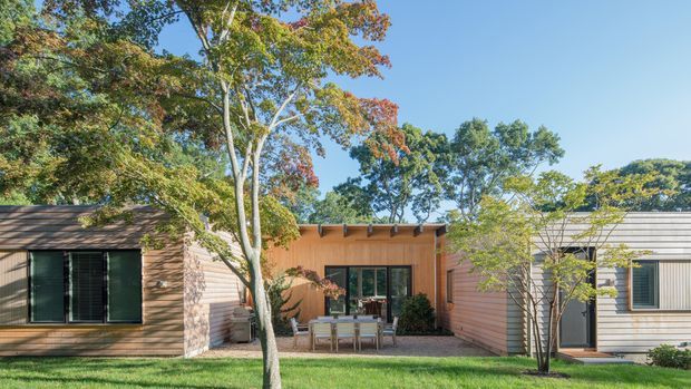 Exterior view of a low, single-storey contemporary residence composed of connected timber-clad volumes around a sheltered central outdoor dining courtyard. The facade features horizontal timber weatherboards, dark-framed glazing, exposed roof-edge beams, and a lighter ribbed metal-clad wing, set within a mature landscaped garden.