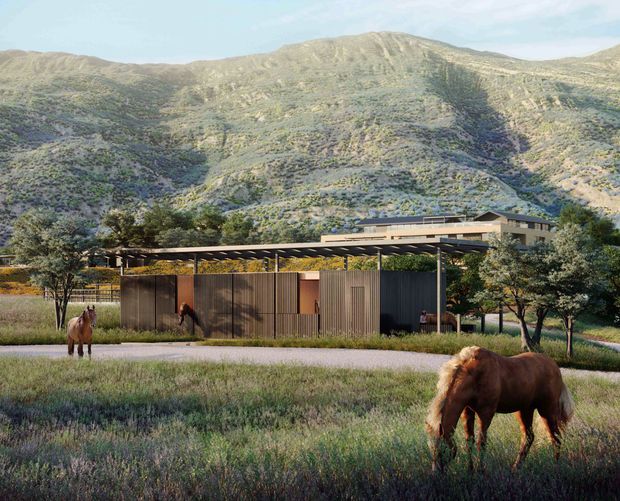 A low-profile contemporary building set within a rugged mountainous landscape, viewed from an open grassy field with horses in the foreground. The structure features dark vertical cladding, broad horizontal roof planes and deep covered outdoor areas, with a larger glazed or dark-roofed volume behind it. Native-looking vegetation and steep rocky hills dominate the setting.