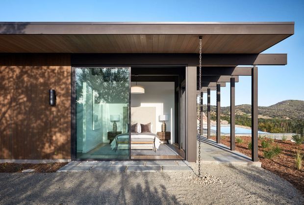 Exterior view of a low, modern pavilion-like dwelling with broad flat projecting roof planes, dark timber-clad walls, extensive sliding glass, and a covered outdoor walkway supported by slender posts. A bedroom is visible through the glazing, while the surrounding arid landscaped site and hills form the background.
