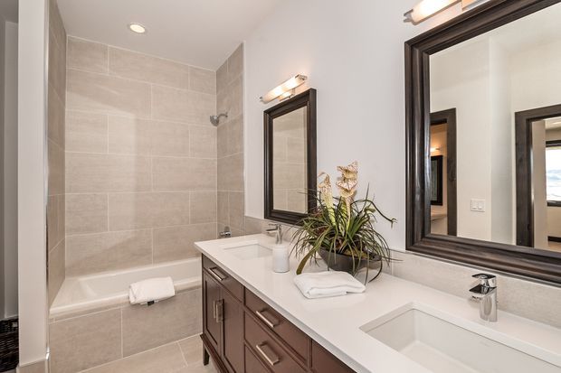 Bright, contemporary bathroom with a long double vanity, white integrated basins, dark wood cabinetry, large dark-framed mirrors, and a tiled bathtub/shower surround. The room uses light beige-grey wall and floor tiles, recessed ceiling lighting, and polished chrome tapware, with a small plant adding natural detail.