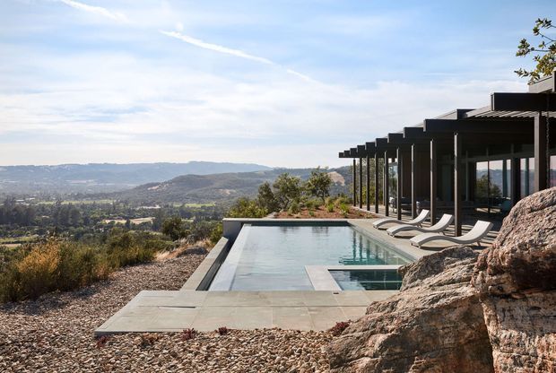 A contemporary hillside residence with a long narrow infinity-edge swimming pool overlooking a broad valley. The pool terrace is finished in large pale stone or concrete slabs and gravel, with several sculptural loungers beside dark-framed floor-to-ceiling glazing. A strongly projecting roof and repeated dark vertical posts create a shaded outdoor edge, while exposed rock outcrops integrate the house into the sloping site.