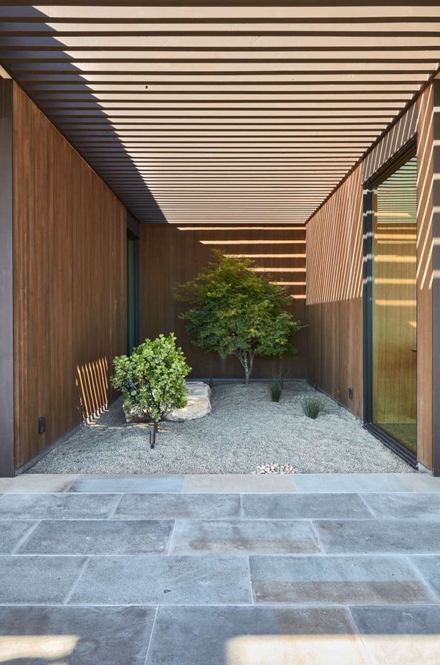 A narrow, sheltered courtyard or passage framed by warm vertical timber-clad walls and a slatted timber overhead canopy. The space contains gravel landscaping, a small ornamental tree, shrubs, grasses, and stone seating, with full-height glazed openings on the right. Large rectangular grey stone paving extends across the foreground. Strong sunlight creates rhythmic striped shadows across the walls, glazing, and ground.