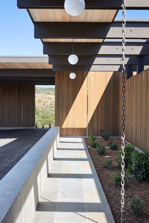 A sunlit covered exterior walkway or courtyard passage beside a modern home. The space features dark exposed structural beams and overhanging flat roof planes, warm vertical timber wall cladding, pale rectangular stone or concrete paving, a low concrete planter wall, and a prominent metal rain chain. Native-style planting lines the walkway, with a dry landscaped hillside visible beyond the opening. Spherical pendant lights hang beneath the covered roof, creating a refined indoor-outdoor architectural composition.