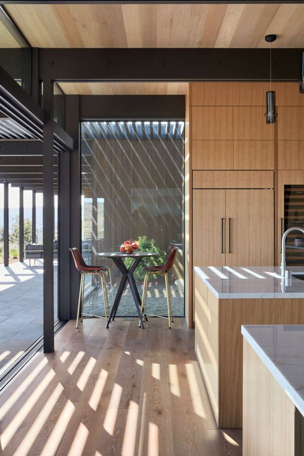 A bright, contemporary kitchen interior with warm vertical timber cabinetry and matching timber ceiling panels. The kitchen includes a pale stone benchtop, integrated appliances, dark-framed floor-to-ceiling glazing, and a compact dining nook with two bar stools and a small table. Strong linear sunlight and shadows enter through the glazed opening and overhead slatted structure, while dark structural framing contrasts with the light wood finishes.