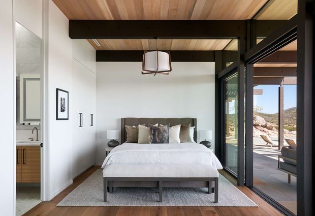 A bright, contemporary bedroom with a neatly made bed, upholstered headboard and bench, framed artwork, and a neutral rug. The room has white walls, warm timber ceiling boards, exposed dark structural beams, and expansive black-framed glazing opening to an outdoor terrace and rocky landscape. A bathroom with timber vanity is partially visible through the opening on the left.
