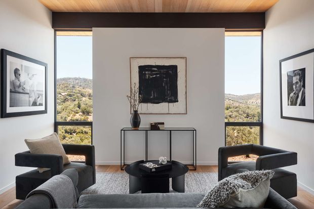 Contemporary living room with a large grey sectional sofa, two dark upholstered lounge chairs, a round black coffee table, and a slim console beneath abstract artwork. Floor-to-ceiling windows frame a hilly landscape, while a warm timber-lined ceiling and dark structural beams add contrast.