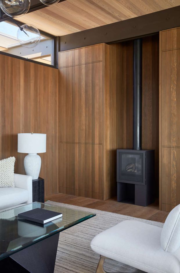 A refined living room with extensive warm timber wall panelling and matching timber ceiling, integrated flush cabinetry, and a freestanding black wood-burning stove with exposed flue. White upholstered seating, a glass coffee table, neutral rug, and sculptural pendant lighting create a warm contemporary interior. High-level glazing brings in natural light.