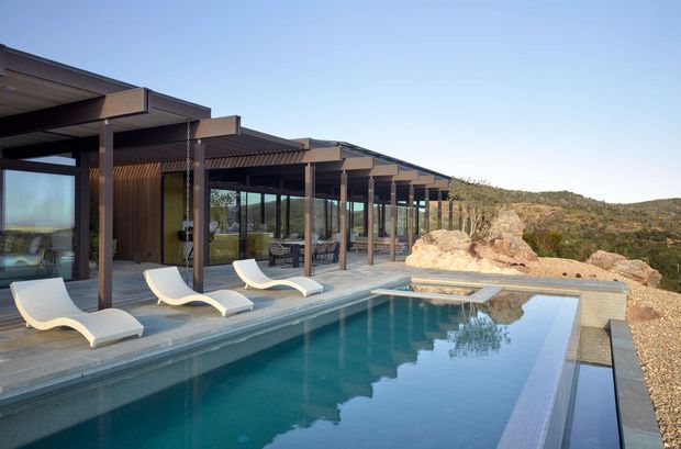 Exterior view of a contemporary single-storey pavilion-style residence with extensive glazing, deep covered outdoor circulation, and a long rectangular swimming pool. White sculptural loungers sit on the pool terrace, while exposed dark structural posts, timber-toned wall cladding, and layered low-pitched roof planes define the building. The home is set against a rugged, dry hillside landscape with natural rock outcrops.