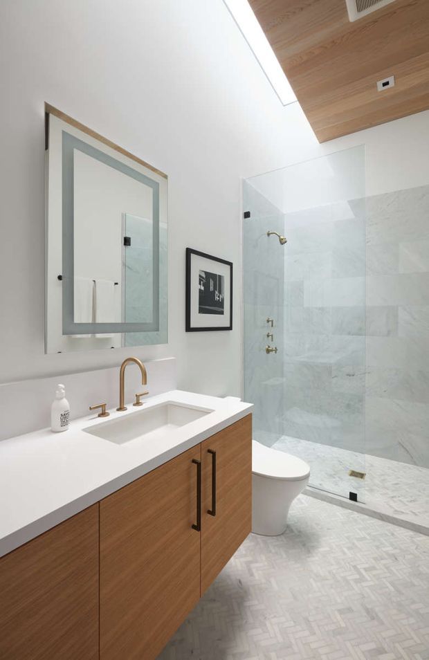 Bright contemporary bathroom featuring a floating timber vanity with white integrated benchtop and rectangular undermount basin, brushed brass tapware, a large framed mirror, white walls, pale marble-look tiled shower walls, and light herringbone floor tiling. The walk-in shower has a frameless glass screen and brass fixtures, with a white toilet positioned beside the vanity. A timber-lined ceiling adds warmth.