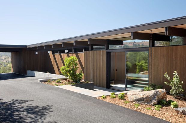 Single-storey contemporary residential building viewed from the driveway, with dark vertical timber cladding, a low-slung shallow-pitched roof, deep roof overhangs, clerestory glazing, and large glazed entry doors. The building is integrated into a landscaped site with exposed rock, gravel planting beds, and distant hills visible beyond.