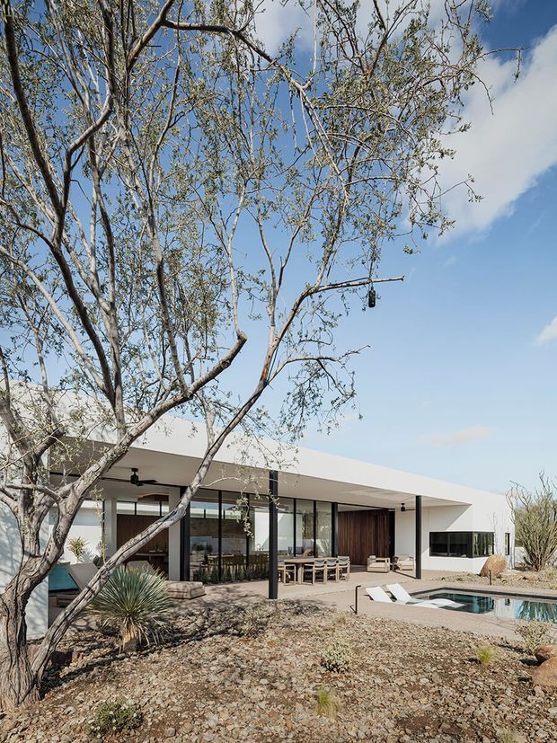 Single-storey contemporary residence with a low, flat roof and extensive floor-to-ceiling glazing, viewed from a dry landscaped garden. The home has crisp white exterior planes, dark slender window and door frames, a covered outdoor dining terrace, timber-clad entry volume, and a swimming pool in the foreground. Sparse desert planting and rocky ground surround the house beneath a bright blue sky.