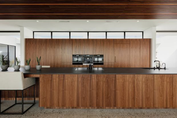 A spacious contemporary kitchen dominated by a long central island and full-height walnut-toned cabinetry. The kitchen features integrated black appliances, a dark benchtop, black-framed glazing, indoor plants, bar seating, and a striking slatted timber ceiling. Terrazzo-like flooring and crisp white walls create a refined, modern interior with warm timber contrast.