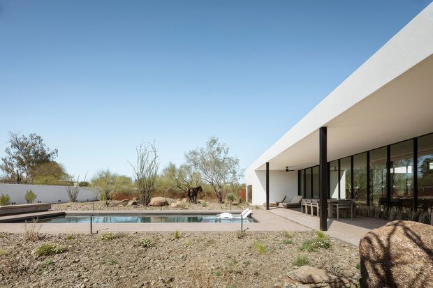 Single-storey modern flat-roof residence viewed from a dry landscaped courtyard, with a long white rendered roof canopy, slender dark supports, extensive floor-to-ceiling glazing, and a rectangular swimming pool in the foreground. The property is set within an arid, desert-like landscape with gravel, sparse drought-tolerant planting, and sculptural rocks. Outdoor seating is visible beneath the covered terrace.