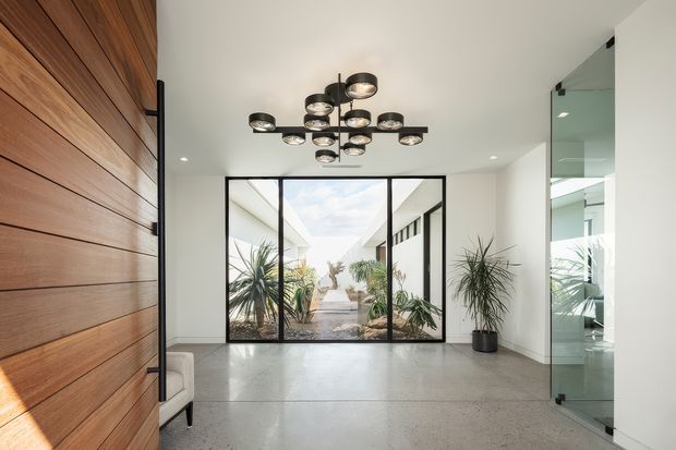 A bright, contemporary entry hall with a polished concrete floor, white walls and ceiling, a full-height timber slat feature wall and black-framed glazed doors opening toward a planted courtyard. A sculptural black multi-light pendant hangs centrally, with a tall indoor plant and reflective glass partition adding visual interest.