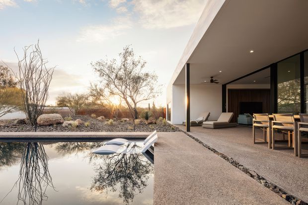A low, modern residence opens onto a tranquil rectangular pool and landscaped courtyard. The image shows a covered outdoor living and dining area with extensive dark-framed glazing, clean white walls, concrete or rendered paving, and minimalist lounge and dining furniture. The surrounding xeriscape garden uses gravel, sculptural rocks, and sparse drought-tolerant planting, with warm sunset light reflected in the still water.