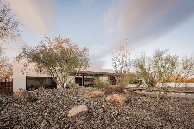 A low, single-storey modern residence set within a deliberately rocky desert landscape. The house has a flat roof, crisp white rendered walls, extensive dark-framed glazing, and a long horizontal composition. Large boulders, gravel, and sparse desert trees and shrubs dominate the foreground, with a paved path or terrace alongside the building.