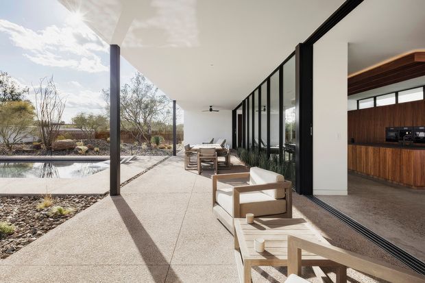 A covered outdoor terrace extends along a glass-fronted modern home, with light concrete paving, timber-framed lounge furniture, and a dining setting. Full-height black-framed glazing opens to a warm timber-lined kitchen interior. A shallow reflecting pool and minimalist desert-style landscaped garden sit beside the terrace, beneath a broad flat roof with slender structural columns.