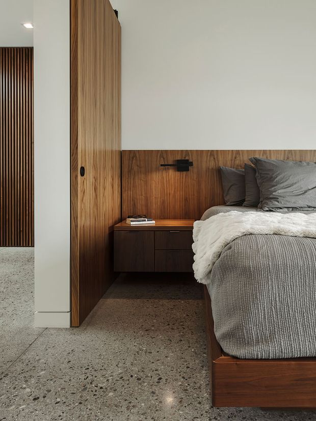 A contemporary bedroom with a low timber bed and integrated dark wood bedside joinery. Vertical timber wall panelling and a timber-clad door create a warm feature wall against smooth white walls. The room has polished exposed aggregate or terrazzo-like concrete flooring, charcoal-grey bedding, a black wall-mounted reading light, and a minimalist floating nightstand. A slatted timber screen is visible through the doorway at left.