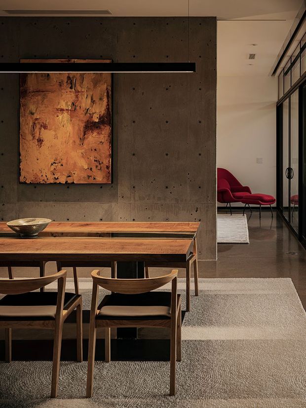 A refined open-plan dining area with a substantial warm timber dining table and sculptural wooden chairs, set against an exposed concrete feature wall. A large abstract artwork and long black linear pendant light anchor the space. Black-framed glazed doors lead toward a lounge area with vivid red lounge chairs. Neutral carpeting, polished dark flooring, and restrained architectural detailing create a sophisticated contemporary interior.