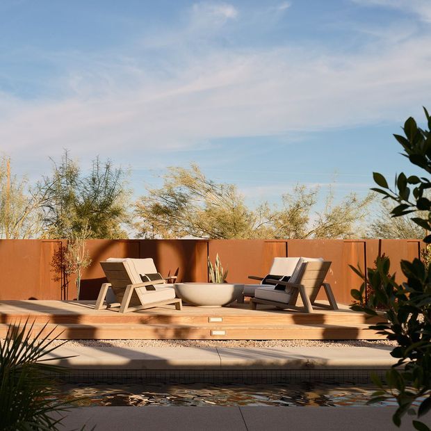 A sunlit outdoor poolside terrace with a warm rust-coloured privacy fence, timber decking, two upholstered lounge chairs, and a low round planter. Desert-like landscaping and filtered sunlight create a relaxed, resort-inspired setting, with the swimming pool visible in the foreground.