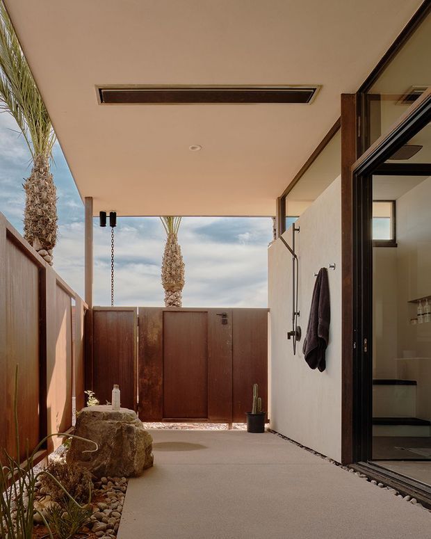 A covered outdoor courtyard or passage beside a home, featuring a smooth rendered ceiling with recessed linear lighting, warm timber privacy fencing and gate, concrete paving, a natural stone feature, pebble landscaping, and desert-style planting. A glazed bathroom or interior space opens directly onto the courtyard at right, with an outdoor shower fixture and towel mounted on the wall. Palm trees and sky are visible beyond the gate.