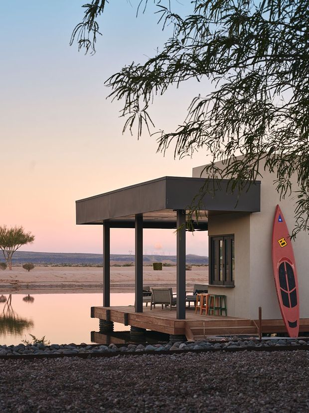 A compact modern lakeside pavilion or cabin at dusk, with a flat-roofed covered deck extending over the rocky shoreline. Dark slender posts support the cantilevered roof, while a warm timber deck, outdoor seating and glimpses of interior glazing create an indoor-outdoor connection. A red paddleboard leans against the pale rendered wall, with calm water and a distant open landscape beyond. Tree branches frame the upper right of the image.