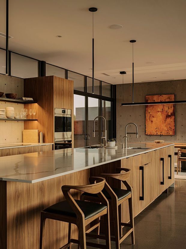 A warm, sophisticated contemporary kitchen with a large central island, integrated wood cabinetry, pale stone benchtop, and seating for several people. The space features black-framed internal glazing, exposed concrete walls, sculptural timber bar stools with dark upholstered seats, and slim black pendant lighting. Built-in appliances and open shelving line the rear cabinetry, while artwork and a dining area are visible beyond the kitchen.