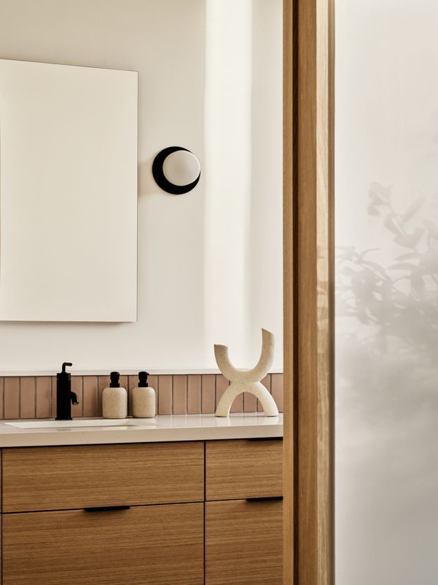 Close-up view of a refined bathroom vanity with warm timber drawer cabinetry, a pale stone or solid-surface benchtop, integrated basin, matte black tapware and wall-mounted fittings. The backsplash features narrow vertically arranged beige-pink tiles, with a large frameless mirror or cabinet above. A timber-framed frosted glass opening or door occupies the right side, softly reflecting foliage-like shadows. The styling is warm, minimal and contemporary with subtle sculptural decor.