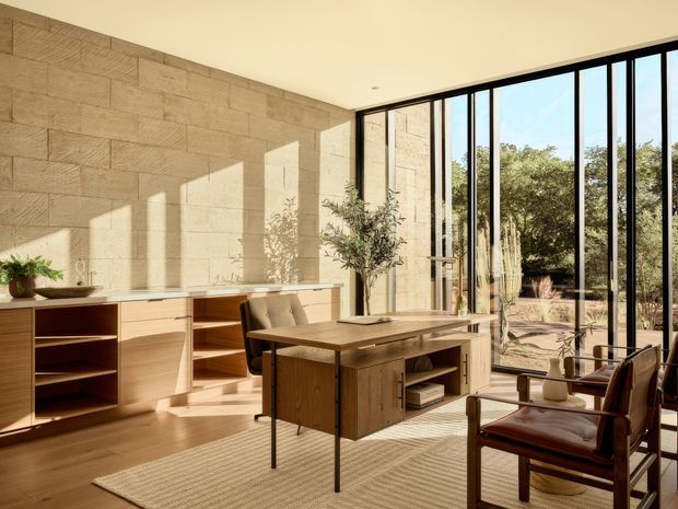 A warm, sunlit modern home office with extensive floor-to-ceiling black-framed glazing overlooking landscaped grounds. The room features a large timber desk, built-in timber cabinetry and shelving along a textured light stone block wall, timber flooring, a woven rug, and a leather lounge chair. Strong sunlight creates geometric shadows across the walls and floor.