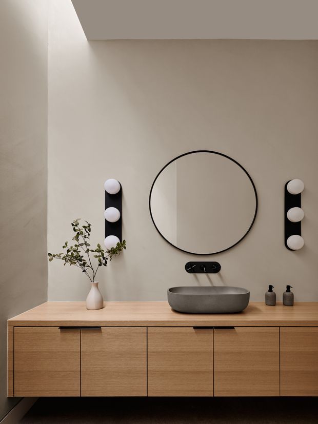 A refined contemporary bathroom vanity with a long floating timber-look cabinet, integrated flat-front drawers and doors, a grey stone vessel basin, wall-mounted black tapware, and a large round black-framed mirror. Two vertical black wall lights with spherical white globes flank the mirror. The warm neutral plaster wall, timber joinery, minimal fittings, and small organic branch arrangement create a calm, understated composition.