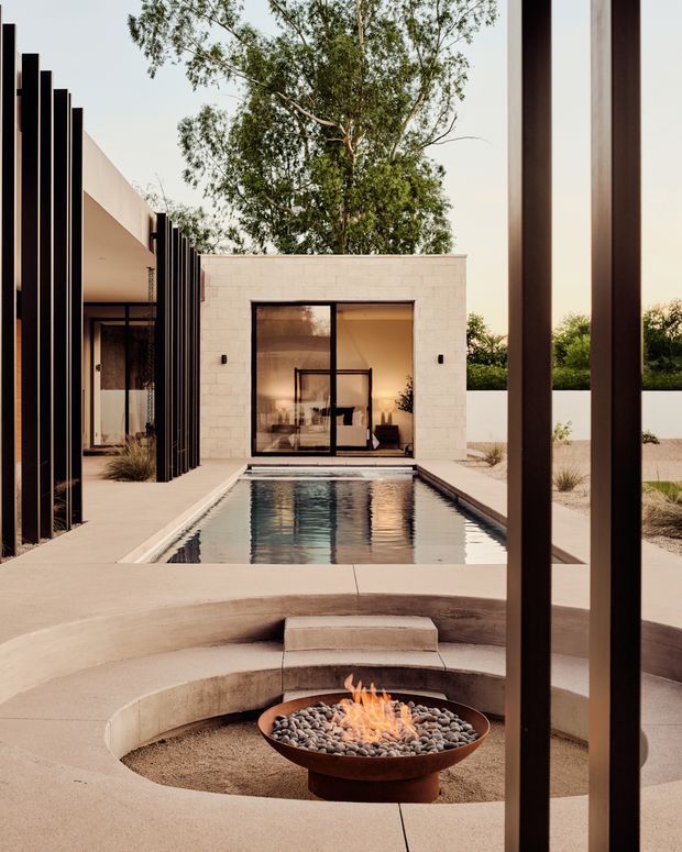 A contemporary courtyard viewed from beneath a covered structure, featuring a narrow rectangular swimming pool, pale masonry walls, dark vertical metal screens, and a circular sunken seating area with a fire bowl in the foreground. Floor-to-ceiling glazing connects the courtyard to the interior, while mature planting softens the geometric architecture.