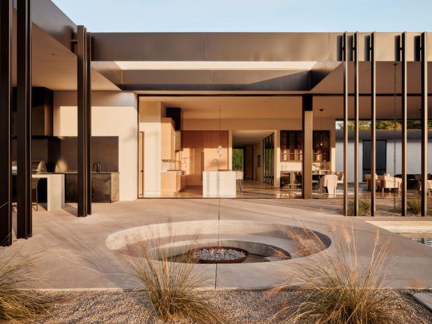 A sophisticated modern courtyard exterior viewed from a landscaped forecourt, with broad concrete paving, a circular recessed fire-pit or water-feature-like element, and ornamental grasses in gravel. The house has a strong rectilinear composition with dark slender vertical metal screens, expansive openings, warm timber-toned interior surfaces, a flat roof, and a central covered threshold connecting to open-plan living spaces. The image is rendered or photographed in warm late-afternoon light.