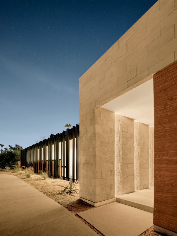 Night-time exterior view of a modern architectural entry or passage. A large pale stone or concrete-block facade forms a deep illuminated opening beside a tall warm timber-clad door. A dark vertical-slat fence extends along the landscaped boundary, with a paved walkway and sparse drought-tolerant planting in the foreground.