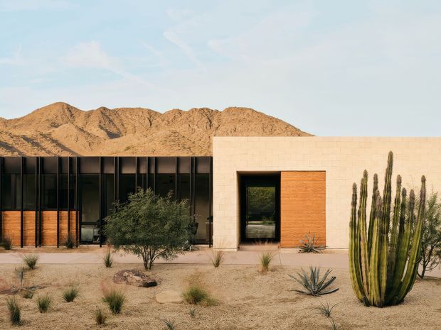 A single-storey modern desert residence with a low, rectilinear form, dark full-height glazing, warm timber screens, and a pale masonry or stucco entry volume. The building sits in a xeric landscaped setting with gravel, agave, grasses, shrubs, and tall columnar cacti, backed by rugged arid mountains under a clear sky.