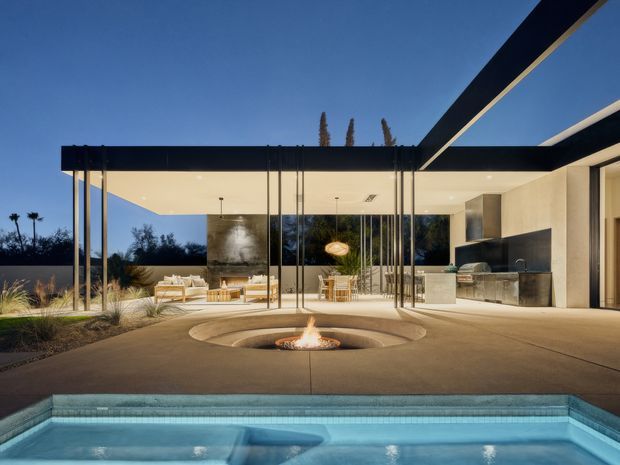 A luxurious modern single-storey pavilion-style home opening onto a pool terrace. The image shows extensive floor-to-ceiling glazing, a strong flat roof with deep black framing, a covered outdoor living area, an open-plan kitchen and dining space, and a sunken circular fire pit in the concrete terrace. The architecture is clean-lined and minimalist, with warm interior lighting and a calm blue-hour atmosphere.