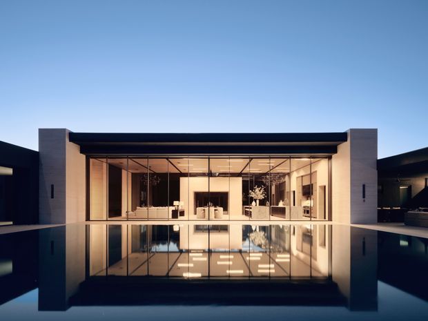 A symmetrical dusk exterior view of a contemporary single-storey pavilion-like residence, featuring a broad floor-to-ceiling glazed facade framed by pale masonry piers and a dark flat roof. The building is reflected in a calm swimming pool in the foreground, with warm interior lighting visible through the glazing.