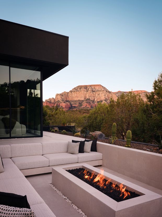 A contemporary outdoor terrace with a built-in L-shaped upholstered seating area surrounding a rectangular gas fire pit. The terrace is enclosed by a low rendered wall and landscaped with desert plants, cacti, and gravel, with dramatic red-rock mountains visible beyond. A dark, floor-to-ceiling glazed corner and projecting flat-roof form frame the left side of the image. The palette is warm and earthy with pale beige surfaces, charcoal accents, and terracotta rock tones.