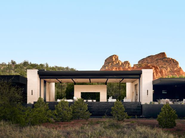 A low, modern single-storey residence set in a dry natural landscape, composed of dark horizontal volumes, pale stone or rendered piers, and a broad open-air covered terrace. The architecture frames views toward dramatic red-rock formations, with extensive glazing, a flat roof, dark retaining walls, and sparse native planting in the foreground.