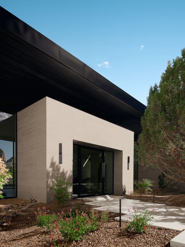Modern residential entrance viewed from outside, featuring a stark rectangular beige masonry or rendered entry volume beneath a broad dark projecting flat roof. The recessed black-framed glazed doorway is approached by a pale concrete or stone path through a minimalist xeriscaped garden with gravel, low shrubs, and slender trees. The composition has strong geometric lines, deep roof overhangs, and a restrained neutral palette.
