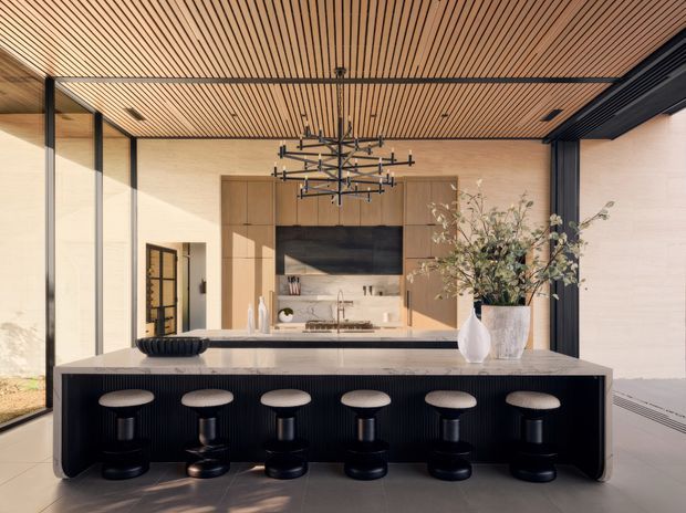 A sophisticated open-plan kitchen viewed straight on, centered on a large dark island with six black bar stools. Warm timber-look cabinetry and a slatted timber ceiling contrast with pale stone or plaster walls, dark-framed floor-to-ceiling glazing, and a dramatic linear sculptural chandelier. The kitchen includes a concealed or integrated cooking zone, sink, pale benchtop, tall cabinetry, and a dark feature splashback. The overall composition is refined, warm, and contemporary.