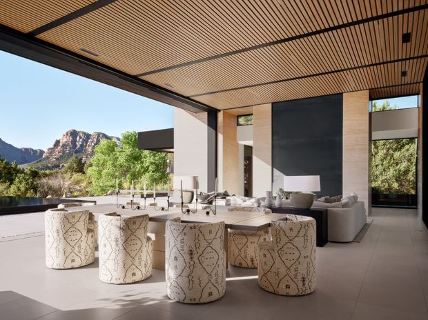 A spacious, open-plan contemporary living and dining area with expansive floor-to-ceiling openings framing a rugged mountain landscape. The room features a slatted timber ceiling with dark structural beams, pale stone or concrete-look floor tiles, warm wood-toned wall panels, and a minimalist kitchen or bar zone in the background. A large dining table is surrounded by distinctive patterned upholstered stools, while neutral lounge seating occupies the adjoining living area. The interior has strong indoor-outdoor integration, abundant daylight, and a refined desert-modern atmosphere.