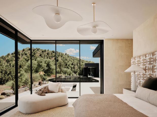A bright contemporary bedroom with expansive black-framed floor-to-ceiling glazing opening toward a lush wooded landscape and a terrace or pool area. The room features a low upholstered bed in warm beige tones, a rounded boucle-style lounge chair, textured neutral textiles, sculptural white ceiling lights, and a geometric patterned feature wall beside the bed. The overall aesthetic is serene, warm, and minimalist with strong indoor-outdoor connection.