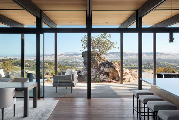 A contemporary open-plan interior with floor-to-ceiling glazing overlooking a wide mountainous valley. The space combines a kitchen island with upholstered bar stools, a dining area, and a lounge chair, under a warm timber-lined ceiling supported by exposed dark structural beams and slender black-framed glazing. Natural rock formations continue into the terrace immediately outside, reinforcing a strong connection to the landscape.