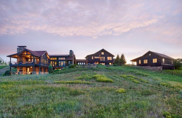 Wide exterior view of a rural multi-building residential compound set in an open grassy landscape at sunset. The complex includes a large timber-clad lodge-like house with balconies and a prominent gabled roof, connected to additional dark-clad gabled buildings. Warm interior lighting glows through the windows, while the surrounding meadow and distant trees reinforce the rural setting.