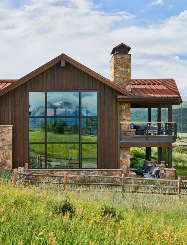 Exterior view of a rustic mountain residence with dark vertical timber cladding, extensive floor-to-ceiling glazing, natural stone walls and chimney, and a rust-red standing-seam metal roof. A raised balcony and covered outdoor seating area overlook a grassy meadow, with dramatic mountains visible through the glazing and beyond the building.