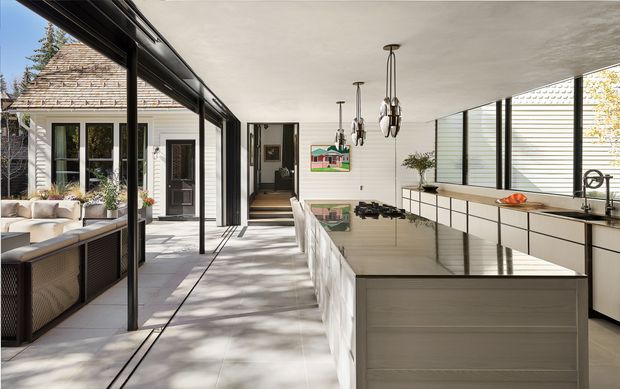 Bright contemporary kitchen opening onto a glazed courtyard or outdoor seating area. The room features a long white island with an integrated dark stone benchtop, pale built-in cabinetry, a wall of horizontal louvres or blinds, dark-framed floor-to-ceiling glazing, and sculptural pendant lights. A timber-shingled exterior and garden seating are visible beyond the glazing.