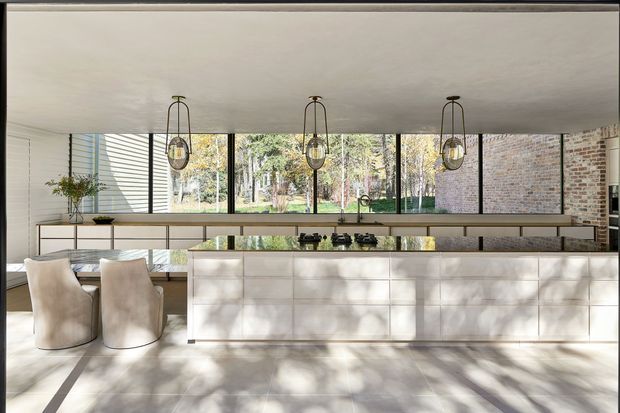 Contemporary open-plan kitchen with a long pale stone or tiled island featuring integrated dark cooktop fixtures, extensive light cabinetry, and a matching rear counter. Three pendant lights hang above the island. Full-height black-framed glazing brings in views of trees, while exposed red-brick walls and strong sunlight add texture and warmth. Two sculptural upholstered bar stools sit at the island.