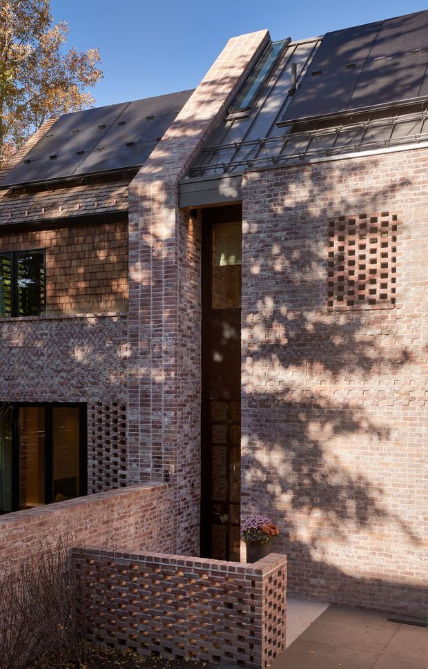 Exterior architectural view of a multi-level brick residence with steeply pitched dark metal roofs, expressed brick walls, narrow vertical glazing, and perforated brick screens forming a sheltered entry courtyard. The composition highlights the layered roof forms, textured masonry, and integrated screened openings in clear daylight.